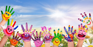 Hände mit bemalten Gesichtern und Blumen auf einem Feld sind in einem Halbkreis angeordnet, mit einem strahlend blauen Himmel und Sonne im Hintergrund.